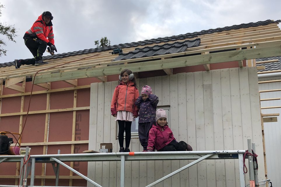 Enebolig under bygging: snekker legger tak mens tre barn står på stillaset